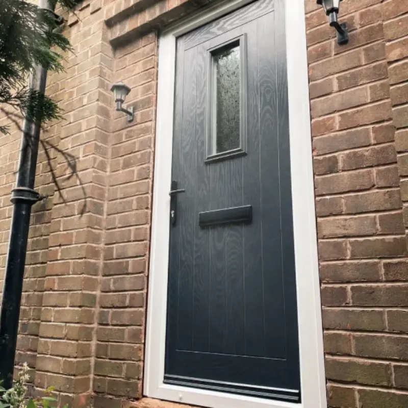 An anthracite composite door with a small obscure-glazed pane and a white frame, set in red brick between two coach lanterns.