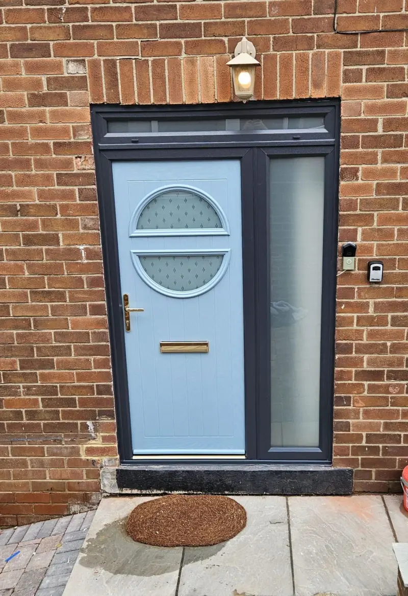 A duck-egg blue composite door with a circular porthole in two patterned halves, brass furniture, an obscure-glazed sidelight and a transom above, in red brick.