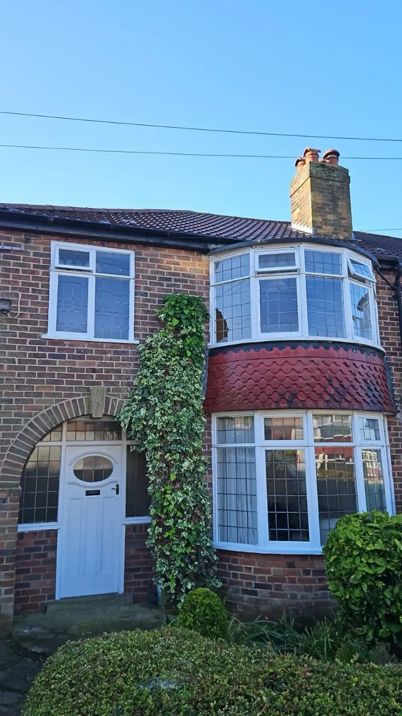 A red-brick semi-detached house with a two-storey bay of white-framed leaded windows, a band of red fishscale clay tiles, ivy climbing the wall, and a white front door with an oval glazed panel set in an arched brick opening.