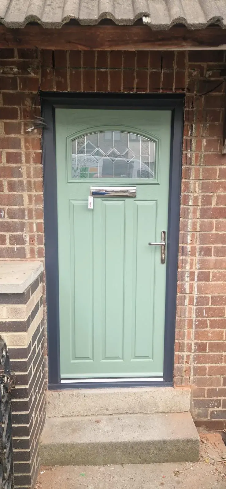 A sage-green composite door with an arched leaded top panel and chrome furniture, in an anthracite frame under a corrugated porch roof.